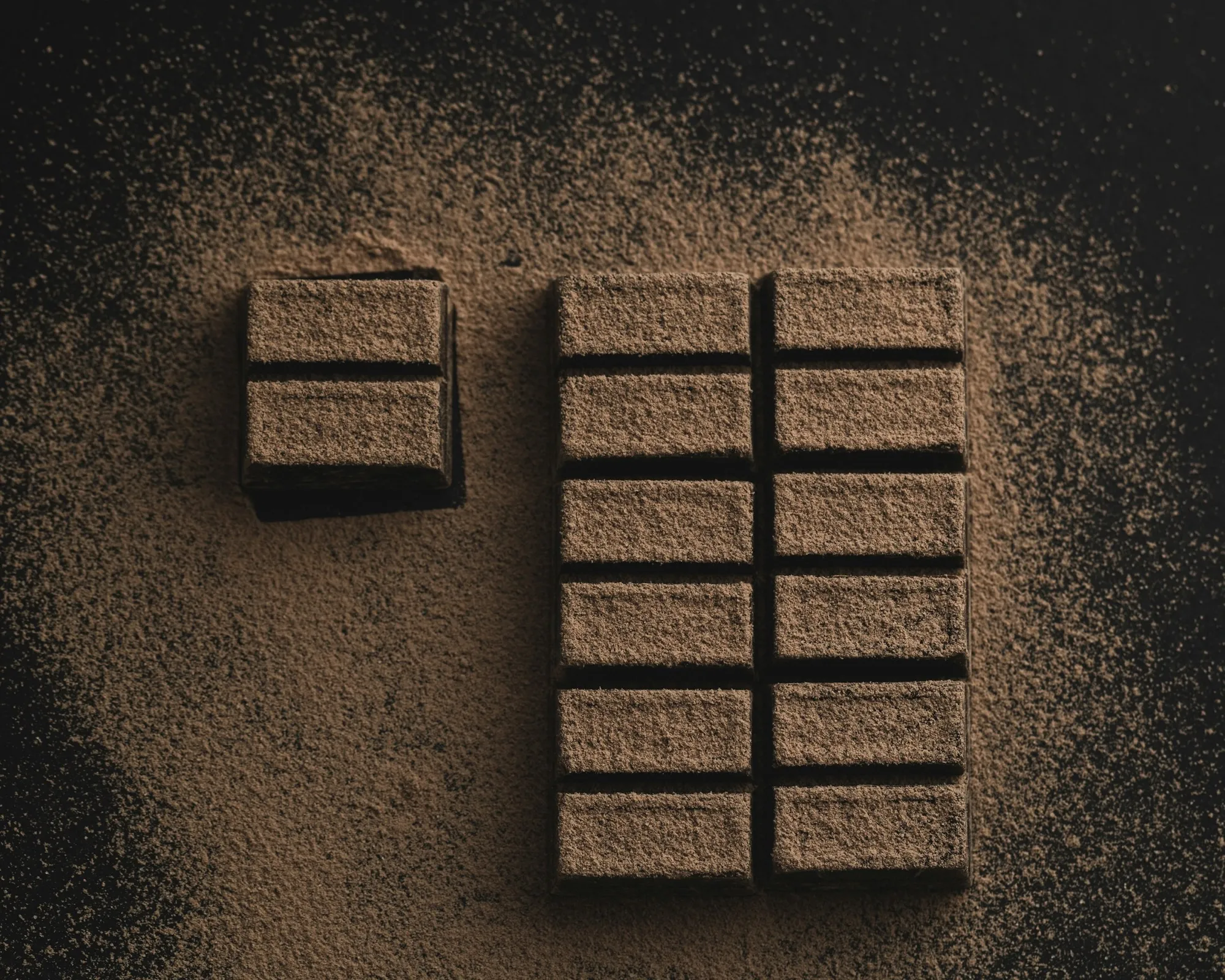 A bar of dark chocolate dusted with cocoa, broken into segments on a dark surface.